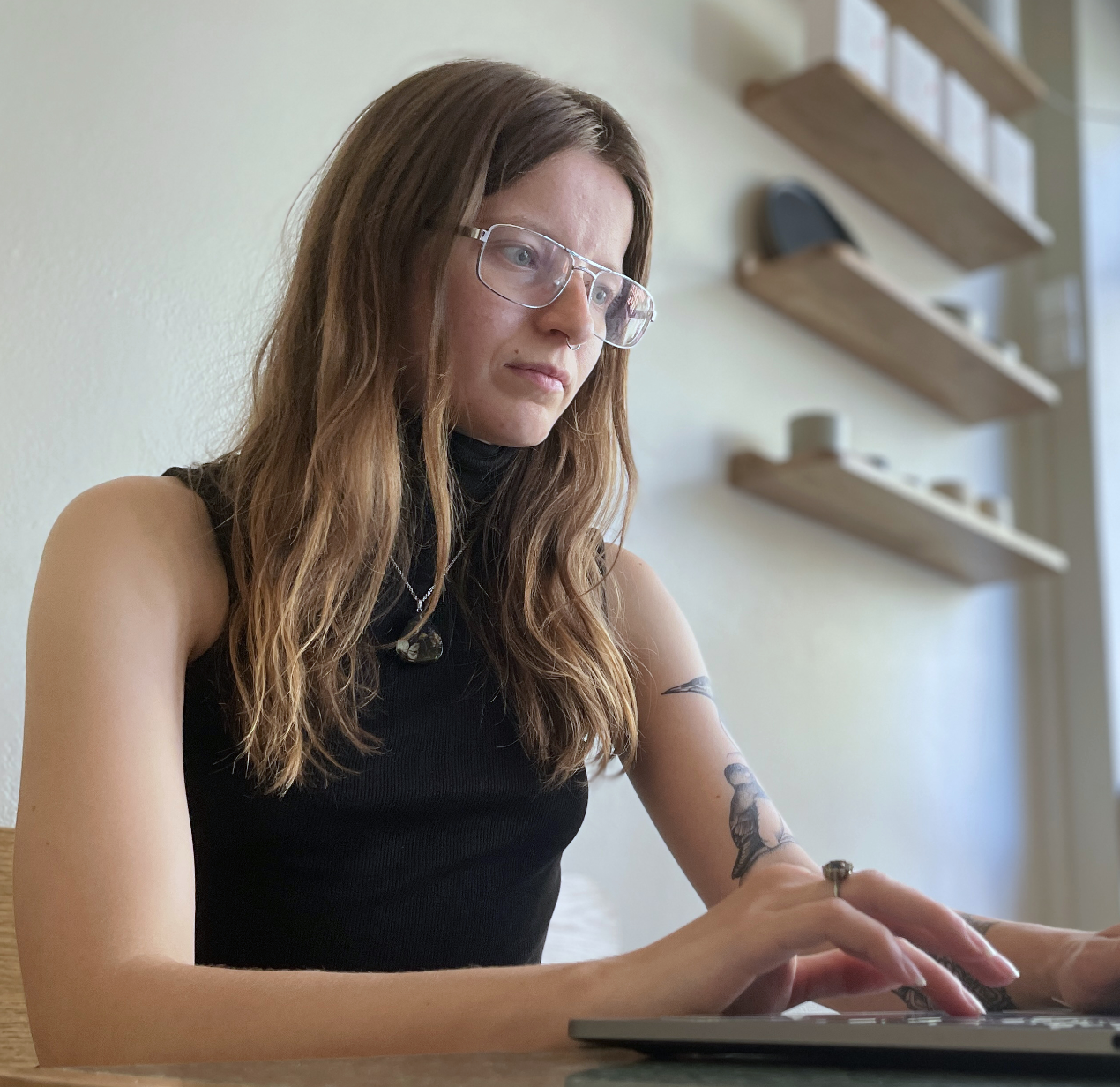 Sarah working, looking deeply focused at their laptop. They have long medium-brown hair, metal rectangular framed glasses, and are wearing a black top.